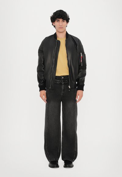 Black leather bomber jacket with ribbed cuffs, yellow t-shirt, and black wide-leg denim jeans. Model wears shiny black shoes.
