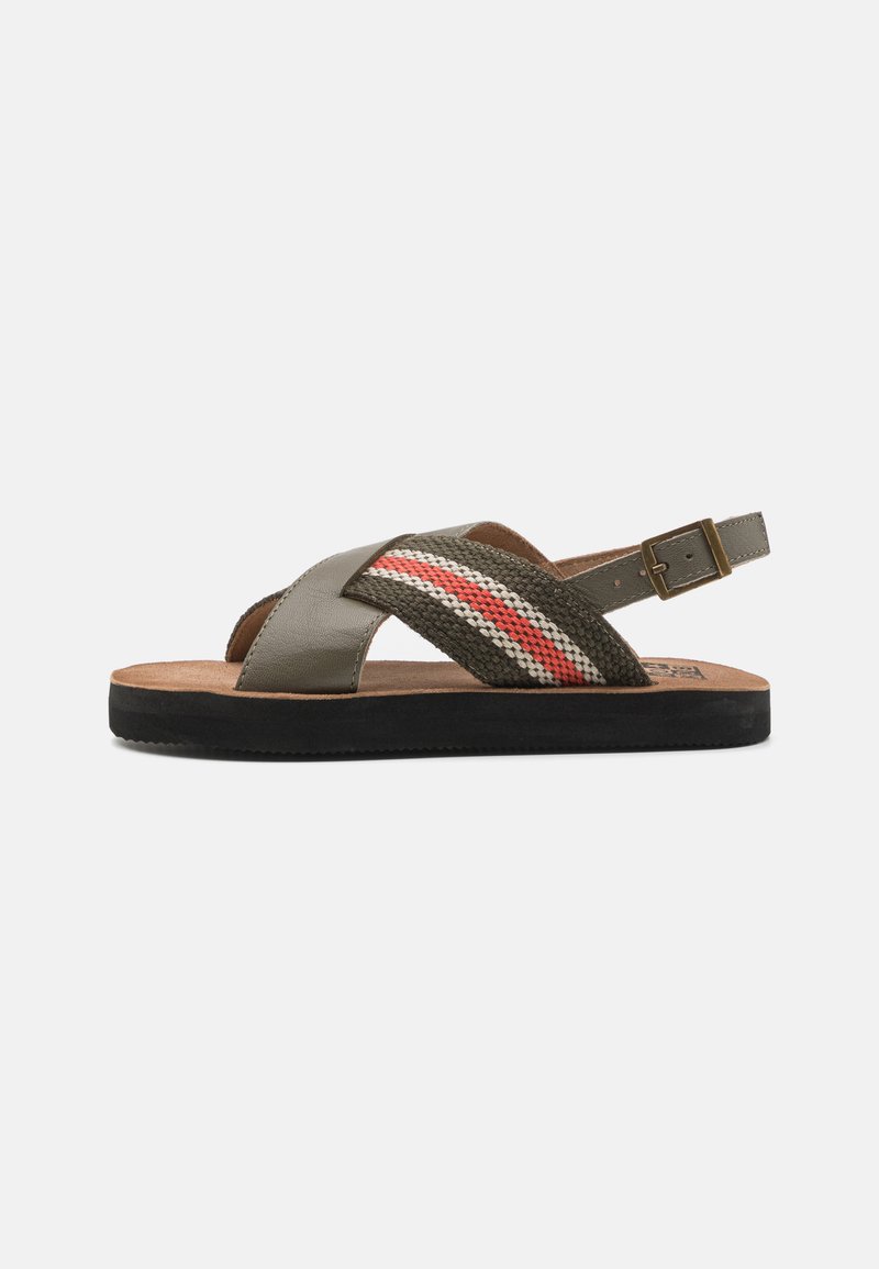 Sandals featuring a gray leather upper with a woven strap in green, red, and cream; adjustable ankle strap; textured, flat black sole.