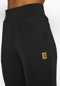 Tuvumā redzami melni sporta bikses ar augstu jostasvietu un mazu daudzkrāsainu Nike logotipa uzlīmi augšstilbā.