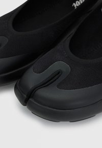 Black split-toe slip-on shoes made of knit fabric with rubber soles and reinforced toe caps on a light background.