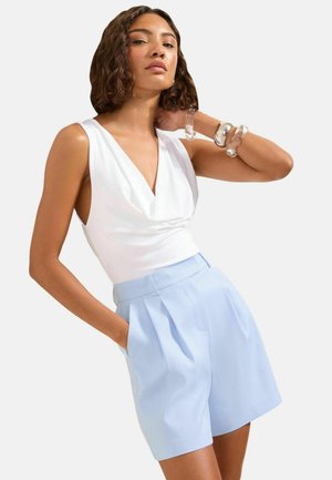 Frau trägt ärmelloses weißes Top und hellblaue, plissierte Shorts mit hoher Taille, posiert mit einer Hand in der Tasche und transparenten Armreifen am Arm.