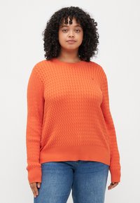 Orange Strickpullover mit strukturiertem Muster, rundem Halsausschnitt und gerippten Bündchen und Saum, kombiniert mit blauen Jeans.
