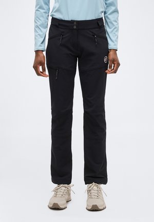 Person wearing black outdoor pants with zippered pockets and light beige hiking shoes, standing against a plain background.