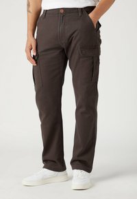 Mies, joka on pukeutunut tummanruskeisiin cargo-housuihin ja valkoisiin lenkkareihin, kädet taskuissa, yksinkertaista vaaleaa taustaa vasten.