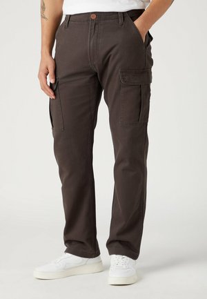 Man in een donkerbruine cargo broek en witte sneakers, met zijn handen in de zakken tegen een effen lichte achtergrond.