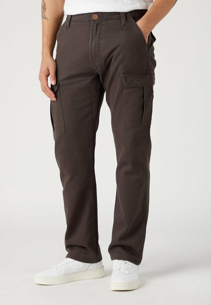 Mies, joka on pukeutunut tummanruskeisiin cargo-housuihin ja valkoisiin lenkkareihin, kädet taskuissa, yksinkertaista vaaleaa taustaa vasten.