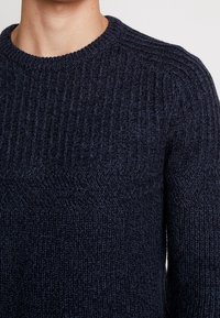 Laivastonsininen neulottu villapaita, jossa on ribbineulos, pyöreä kaula-aukko ja paksu lanka. Neuleessa on vaakasuoria ja kalanruotokuvioita.