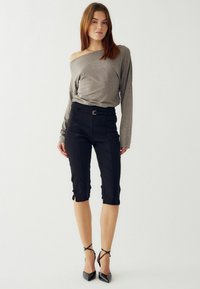 Pelēks gara piedurkne bez plecu tops, melni piegriezti apgriezti bikses ar sānu šķēlumiem, kombinēts ar melniem norādītiem augstpapēžu apaviem.