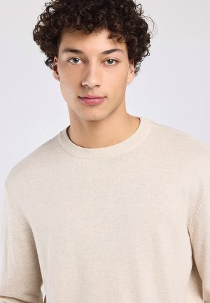 Jonge man met krullend donker haar en lichte ogen, die een beige trui met ronde hals draagt, kijkt recht vooruit tegen een eenvoudige lichte achtergrond.