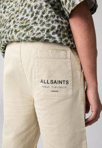 Beiga bomullsbyxor med en bakficka som har en tryckt "ALLSAINTS"-logotyp och text, slät textur och minimalistisk design.