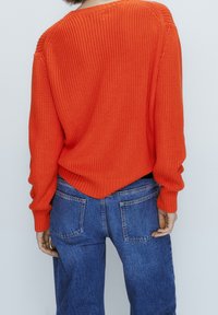 Un pull en tricot orange vif avec une coupe ample, des poignets et un ourlet côtelés, associé à un jean en denim. La texture est tissée avec des coutures visibles.