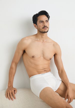 Weiße Baumwoll-Boxershorts mit einem GANT-Logo auf einem weichen Bund. Das Model hat kurzes, dunkles Haar und einen hellen Hautton und sitzt vor einem neutralen Hintergrund.