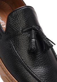 Scarpa slip-on in pelle nera con finitura testurizzata, accentuata da frange e suola contrastante marrone. Elementi di design classici e funzionali.
