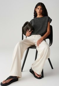 Top a maniche corte con motivo nero, pantaloni larghi crema, sandali neri, tiene una borsa scura e texture. Modella seduta su una sedia nera.