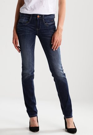 Kobieta ubrana w ciemnoniebieskie jeansy o obcisłym kroju i czarne buty na wysokim obcasie, stojąca na jasnym, jednolitym tle.