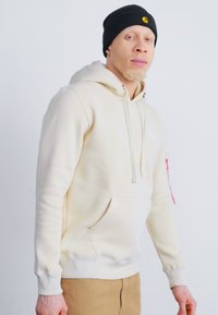 Hoodie crème en mélange de coton, doté d'une poche avant, de cordons de serrage et d'un logo avec des accents rouges. Porté avec un bonnet noir.
