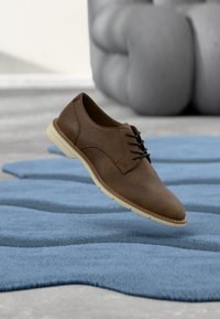 Chaussure à lacets en cuir marron flottant au-dessus d'un tapis bleu texturé avec un canapé gris en arrière-plan flou.