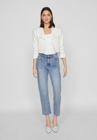 White cropped jacket with a folded collar, worn over a white top. Light blue high-waisted jeans with frayed hem. Pointed white heels.