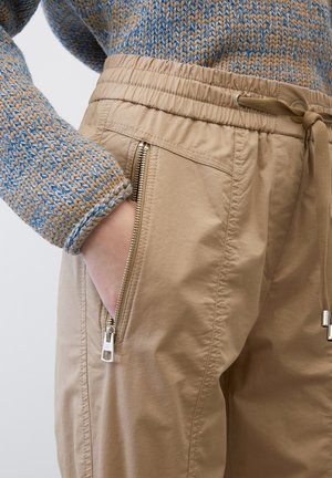 Lichte beige joggers met een elastische tailleband, trekkoordsluiting en ritszakken aan de voorkant. Draag ze met een blauw en bruin gestructureerd sweatshirt.