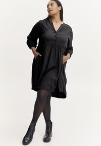 Schwarzes Kleid mit 3/4-Ärmeln, Knopfleiste und geraffter Taille. Verfügt über seitliche Taschen und eine fließende Silhouette. Getragen mit schwarzen Ankle Boots.