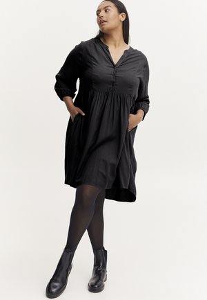 Rochie neagră cu mâneci 3/4, placket cu nasturi și talie strânsă. Prezintă buzunare laterale și o siluetă fluidă. Purtați cu ghete negre până la gleznă.
