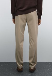 Beige, straight-cut trousers made of smooth fabric, featuring a pleated front, worn with a dark brown sweater, view from the back.