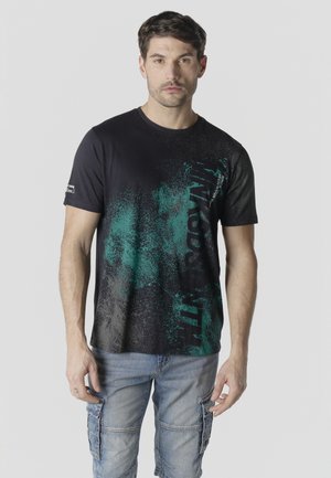Mann trägt schwarzes T-Shirt mit grünem Farbspritzer-Design und Schriftzug, kombiniert mit hellblauen Cargo-Jeans, steht vor einfachem Hintergrund.