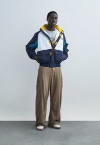 Muž stojaci s rukami v vreckách, oblečený v farebnej bloku bunde, šedom tričku s potlačou, voľných hnedých nohaviciach a bielych teniskách.