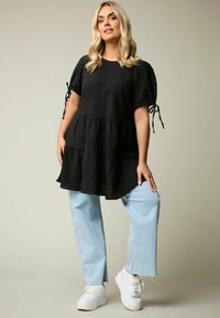 Top nero con maniche a sbuffo in tessuto testurizzato, design a strati e lacci regolabili alle maniche, abbinato a jeans dritti blu chiaro e sneaker bianche.