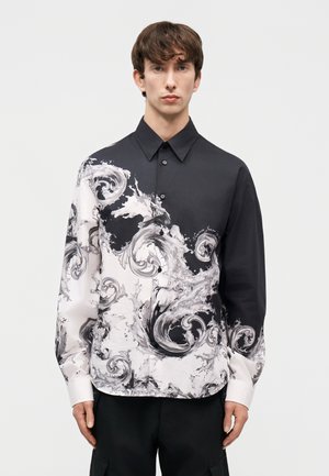 Νέος άνδρας που φοράει ένα μαύρο και άσπρο πουκάμισο με κουμπιά και κυματιστά κυματικά μοτίβα, στέκεται μπροστά σε ένα απλό φωτεινό φόντο.