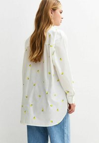 Blouse blanche avec de petites broderies de citrons jaunes, à manches longues et coupe ample. Détail de pli à l'arrière, ourlet arrondi, associée à un jean bleu clair.