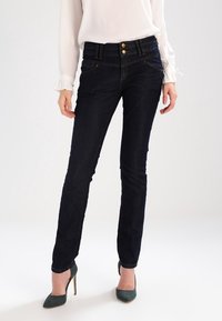 Jeans de denim azul oscuro con corte ajustado, dos bolsillos frontales y cierre de doble botón. Combinados con una blusa de color claro y tacones verdes.