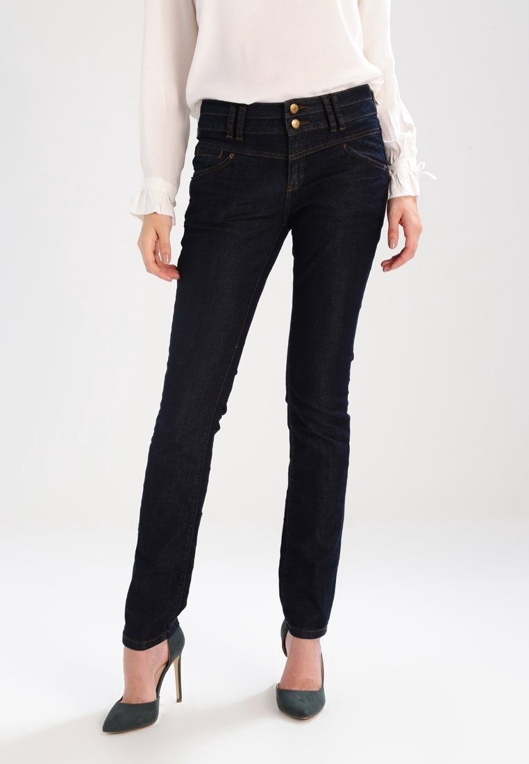 Jeans de denim azul oscuro con corte ajustado, dos bolsillos frontales y cierre de doble botón. Combinados con una blusa de color claro y tacones verdes.