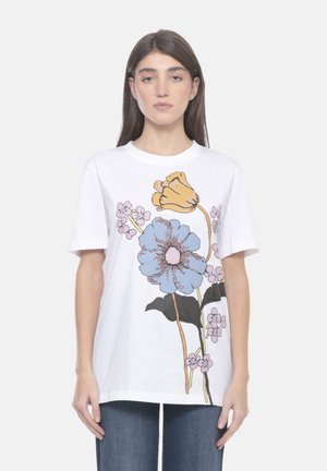 Donna che indossa una maglietta bianca con grande stampa floreale in blu, giallo, rosa e nero, abbinata a jeans blu su uno sfondo neutro.