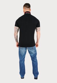 Man viewed from the back, wearing a black polo shirt with a red logo collar and light blue jeans, standing against a plain background.