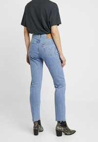 Jeans de mezclilla azul claro con cintura alta, corte recto y dobladillo deshilachado. Cuenta con un parche de cuero marrón y bolsillos traseros con detalles de costura.