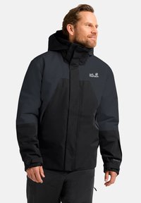 Veste imperméable noire et gris foncé avec capuche, présentant un logo, des poignets élastiques et une fermeture éclair. Convient pour les activités de plein air.