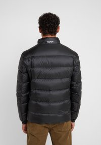 Mann mit lockigem Haar, der eine schwarze Steppjacke mit dem Text "HUGO" am Kragen trägt, kombiniert mit braunen Cordhosen, von hinten gezeigt.