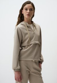 Felpa con cappuccio beige in tessuto testurizzato, con zip frontale e cordino. Vestibilità ampia con polsini elasticizzati. Abbinata a pantaloni coordinati.