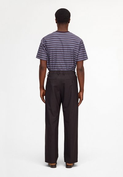 Man standing facing away wearing a blue and gray striped short-sleeve shirt tucked into loose black pinstripe trousers.
