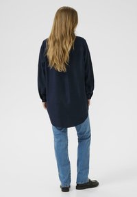 Chemise bleu marine à manches longues avec une coupe droite, dos boutonné et coupe décontractée, associée à un jean en denim bleu clair et des chaussures noires.