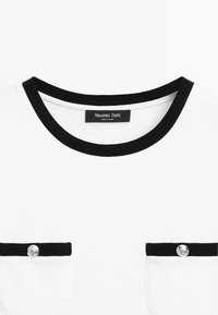 Witte top met een zwarte ribgebreide halslijn en twee borstzakken, met zilveren knoopaccenten. Gemaakt van een zachte, lichte stof.