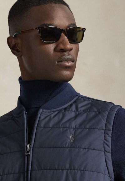 Veste matelassée marine avec fermeture éclair et logo brodé, assortie à des lunettes de soleil foncées avec des verres teintés marron et des accents contrastants sur les branches.