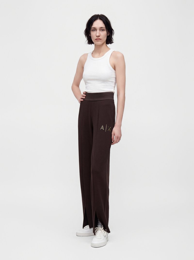 Jeune femme aux cheveux courts et foncés portant un haut blanc sans manches, un pantalon large foncé avec le logo A|X et des baskets blanches, debout devant un fond blanc.