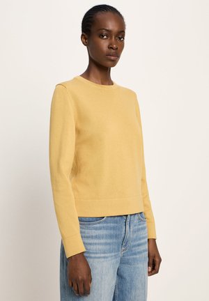 Frau mit geflochtenem Haar, die einen schlichten gelben Pullover mit langen Ärmeln und hellblauen Jeans trägt, steht vor einem neutralen Hintergrund.