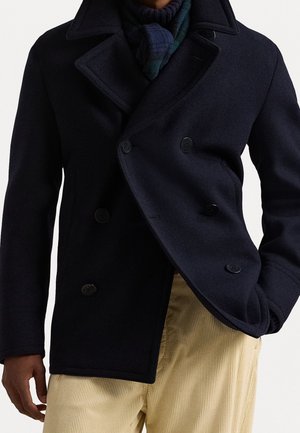 Persoon die een donkerblauwe dubbelzijdige pea coat draagt, een donkere sjaal en lichtbeige corduroy broek, met één hand in de zak.