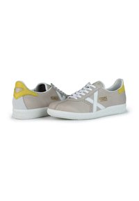 Zapatillas de cuero beige con acentos blancos, que cuentan con una suela blanca texturizada y una sección de talón amarilla. Incluyen cierre con cordones y un diseño perforado.