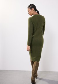 Vestido de punto de color verde oliva con manga larga, corte ajustado, patrón texturizado y longitud media hasta la pantorrilla, combinado con botas marrones por encima de la rodilla.