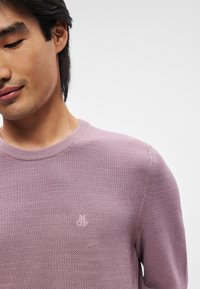 Lyse lilla sweater med struktureret strik, rund halsudskæring og diskret logo på brystet. Glat materiale med ribbet krave og manchetter.