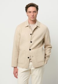Giacca in lana beige con colletto, tasche frontali, sei bottoni neri e taglio dritto. Indossata sopra una maglia chiara e pantaloni.